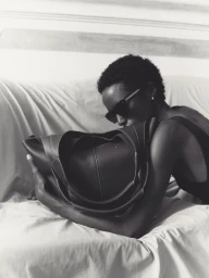 Model holding a dark leather tote bag while resting on a white sofa in a black and white fashion portrait.
