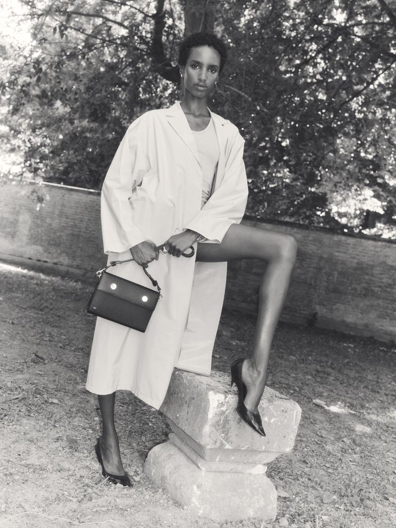 Model posing with a handbag outdoors wearing a light coat and dark heels, shot in monochrome.