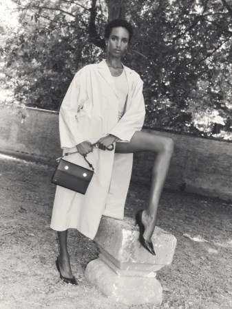 Model posing with a handbag outdoors wearing a light coat and dark heels, shot in monochrome.