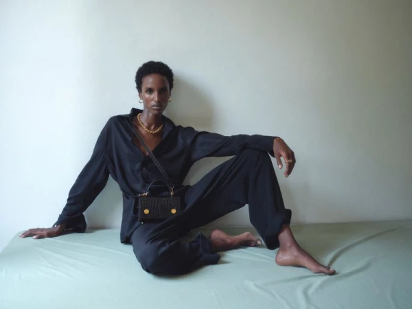 Model Bibi Abdulkadir seated wearing a black shirt and trousers with a black handbag against a pale green and white background