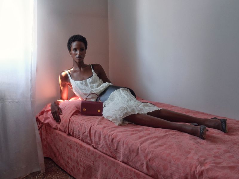 Model in white dress reclining on a pink patterned bed with a dark red handbag in a sunlit room.