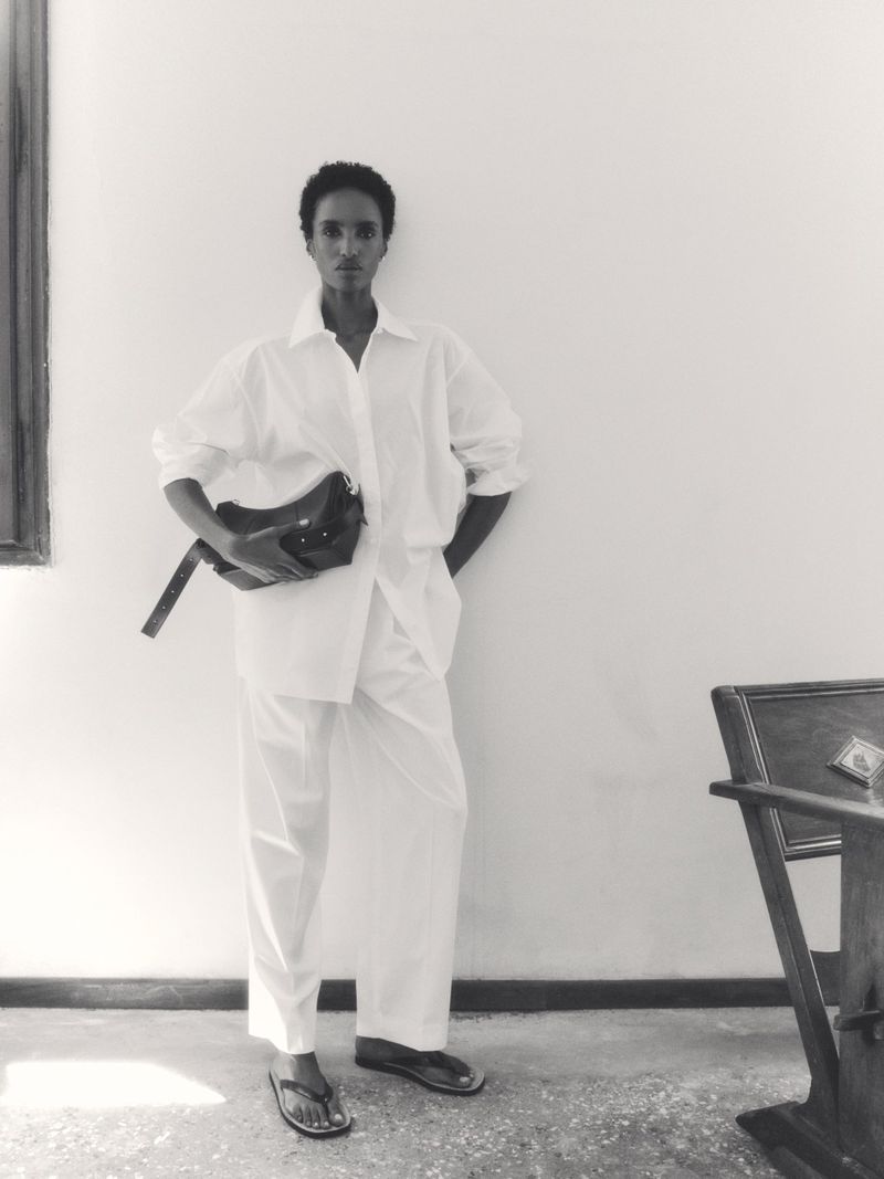 Model Bibi Abdulkadir wearing an all-white outfit and holding a dark handbag against a plain wall.