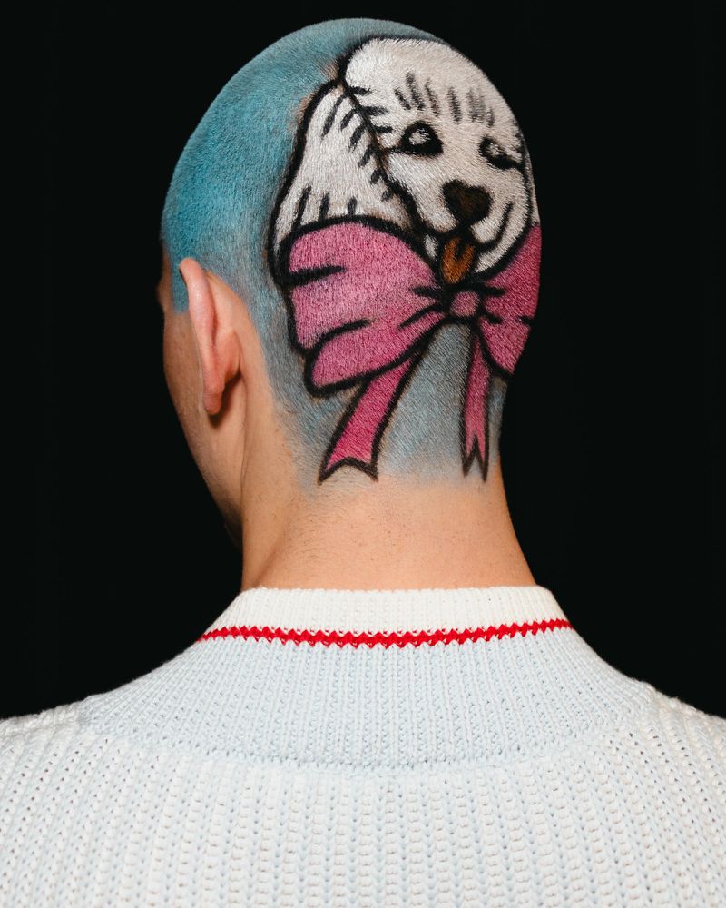 Person with blue-dyed hair and a whimsical puppy and pink bow design shaved into the back, wearing a textured white sweater.