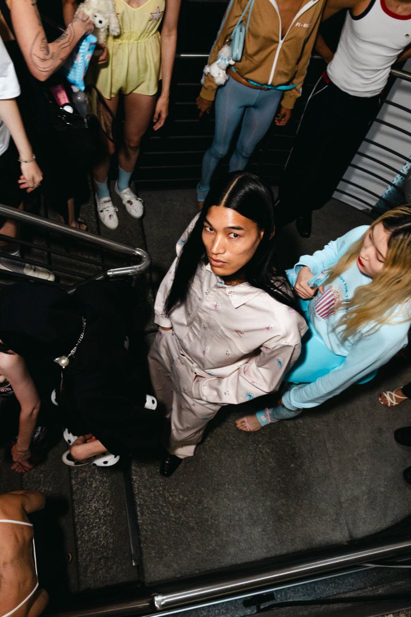 Overhead shot of an Asian woman in pale pink pajamas, surrounded by people in casual streetwear on concrete steps.