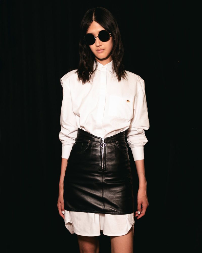 Woman in a white button-down shirt and black leather mini skirt, wearing sunglasses, standing against a dark background in a studio shot.