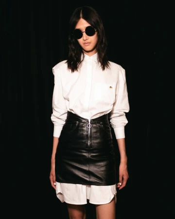 Woman in a white button-down shirt and black leather mini skirt, wearing sunglasses, standing against a dark background in a studio shot.