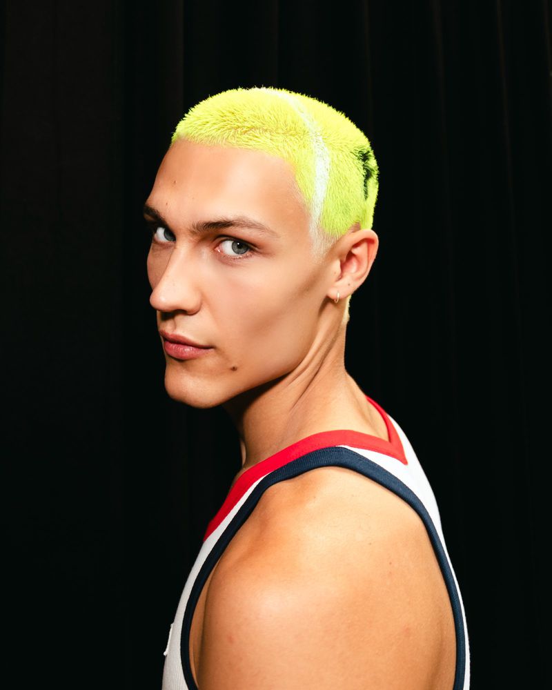 Close-up studio portrait of a young adult man with bright yellow dyed hair. He is wearing a white tank top with red and blue trim.