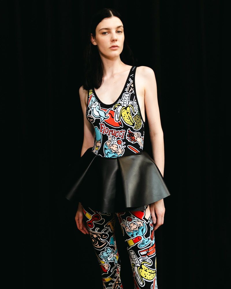 Fashionable woman in printed bodysuit with cartoon graphics and black peplum stands against a black backdrop in a studio setting.