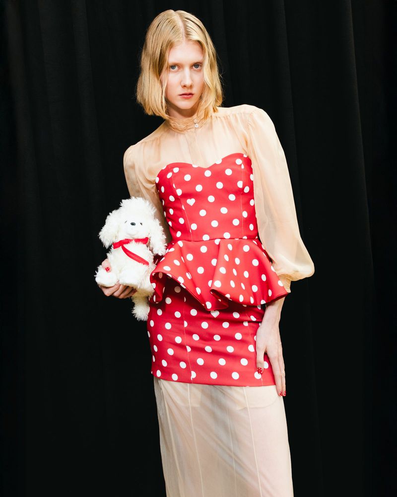 Young woman poses in a red-and-white polka-dot dress, holding a white stuffed dog toy against a dark background in a fashion shot.