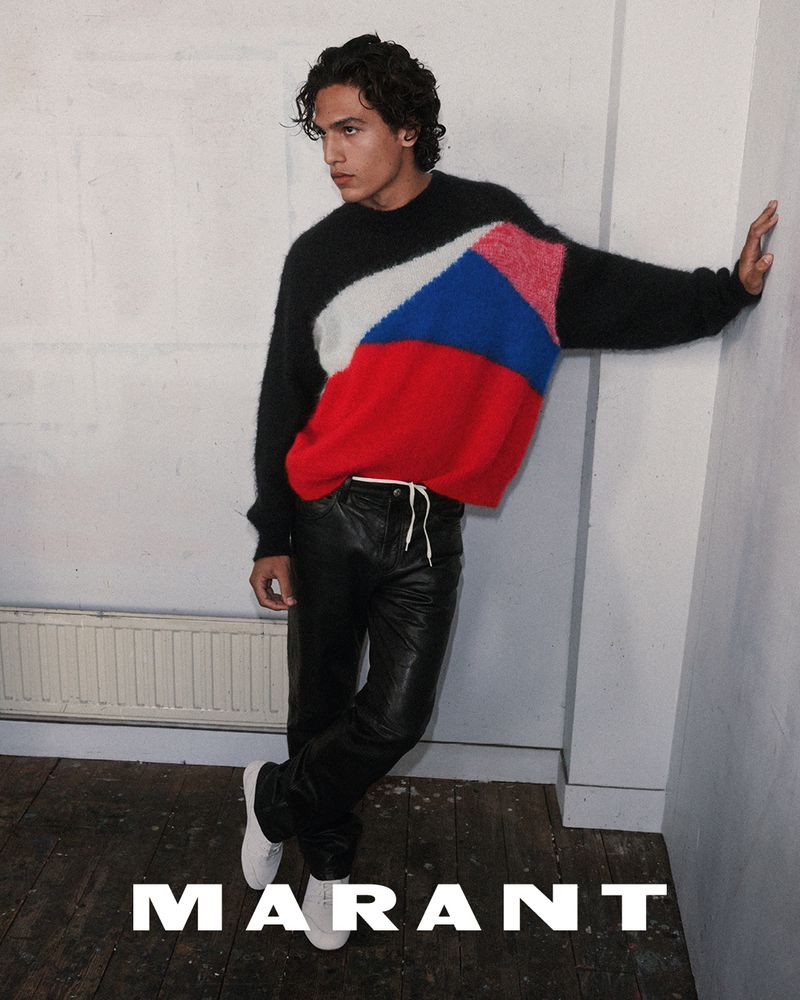 Model wearing colourblocked sweater and black trousers leaning against a white wall with the Marant logo at the bottom