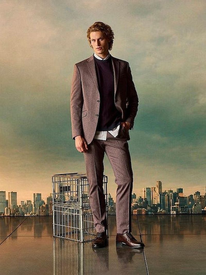 Man wearing a brown suit and dark sweater standing before a city skyline backdrop for Kenneth Cole FW 26 campaign.