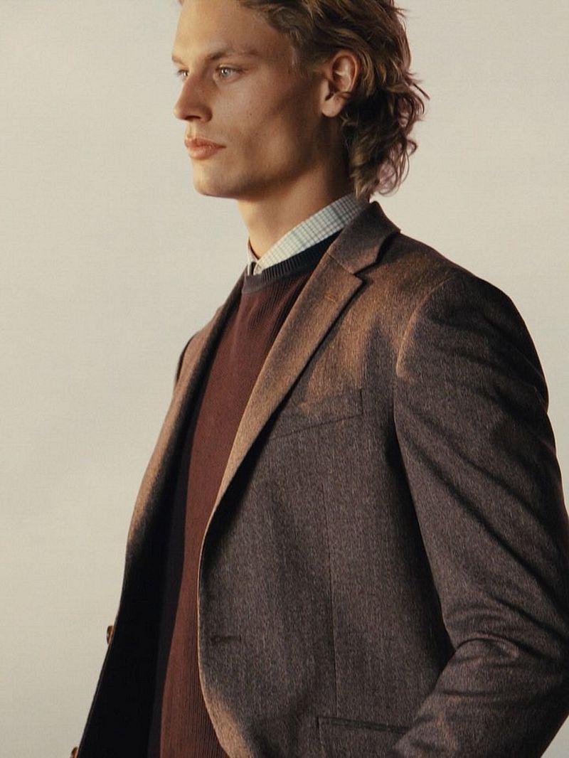 Profile portrait of a male model wearing a brown blazer, dark sweater, and patterned collared shirt against a neutral background.