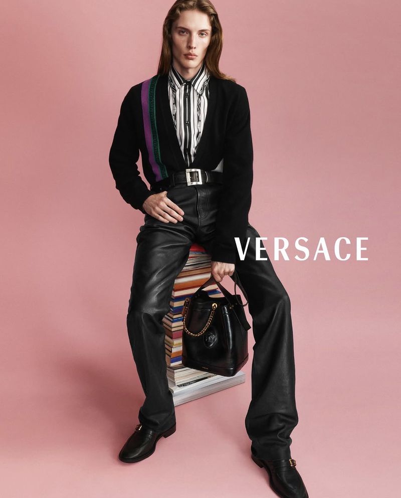 Model Wiley Rhoads seated on a stack of books against a pink backdrop for Versace, wearing a black cardigan and leather pants.