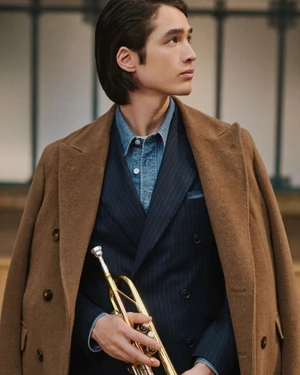 Model wearing a brown coat and pinstripe suit holding a trumpet for the Canali FW26 collection campaign.