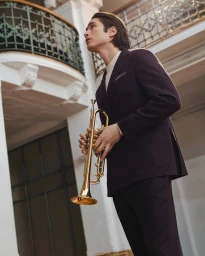 Man in a dark suit holding a golden trumpet inside a classical interior with a balcony railing