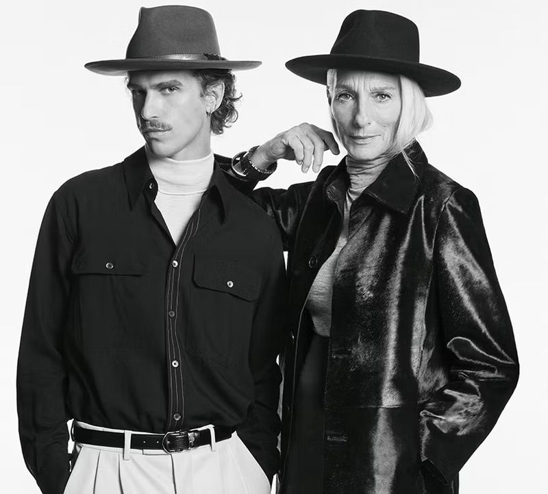 Monochrome portrait of two models wearing wide-brimmed hats and dark coats against a bright white background.