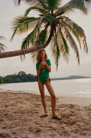 Model in green swimsuit holding a coconut standing on a sandy beach beneath a leaning palm tree