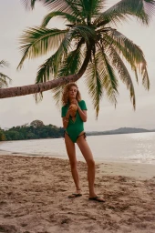 Model in green swimsuit holding a coconut standing on a sandy beach beneath a leaning palm tree