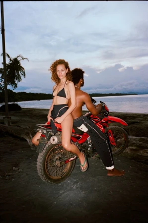 Model Gemma Janes in a black bikini sitting on a red dirt bike on a dark sandy beach at dusk, photographed by Diana Louise Bartlett.