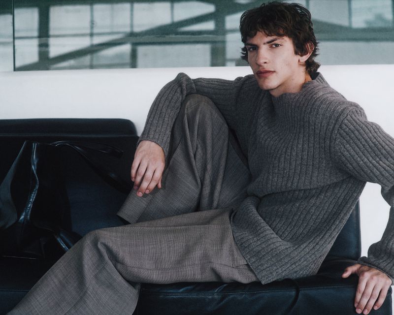 A young male model wearing a grey ribbed sweater and wide-leg trousers seated on a black leather couch indoors.
