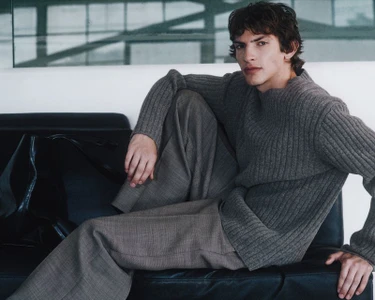A young male model wearing a grey ribbed sweater and wide-leg trousers seated on a black leather couch indoors.