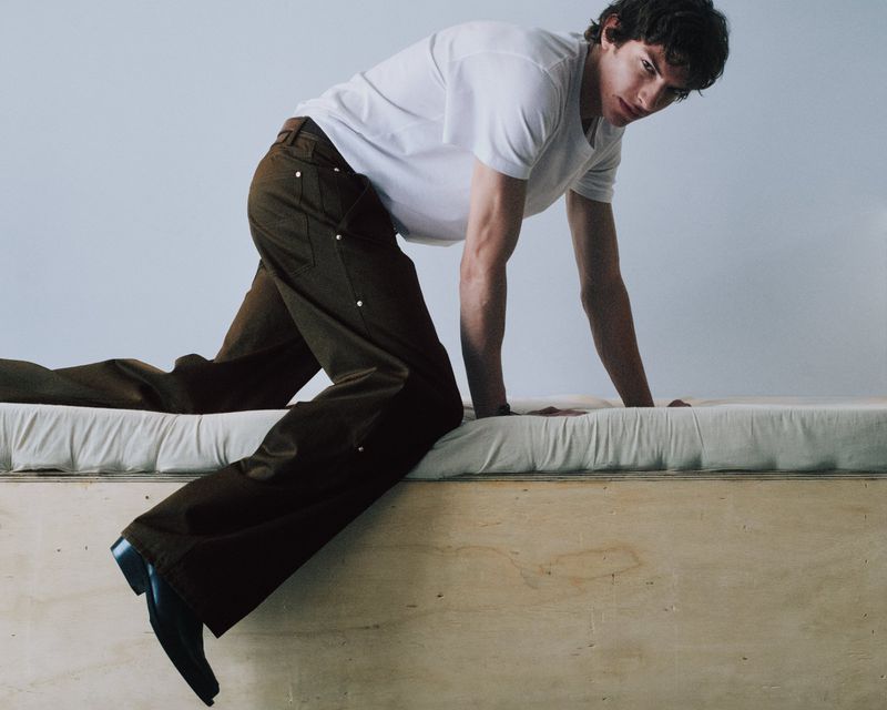 Model in white t-shirt and brown trousers crawling on a wooden platform against a plain grey background
