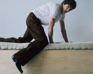 Model in white t-shirt and brown trousers crawling on a wooden platform against a plain grey background