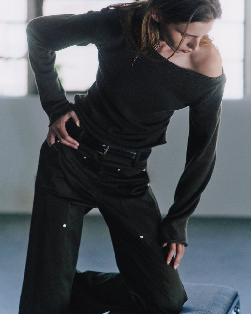 Model in a dark off-the-shoulder sweater and dark trousers posing in an indoor setting.