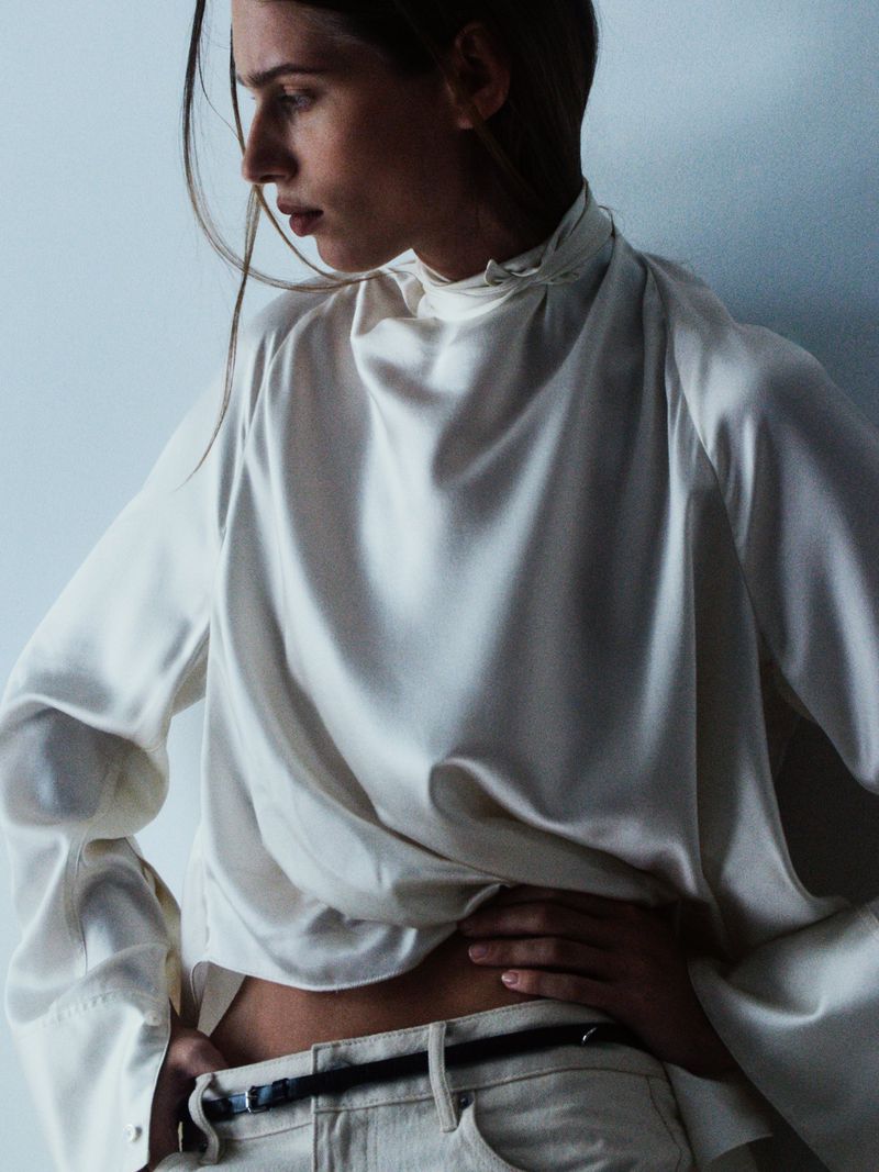 Model wearing a white satin high-neck top and light-colored trousers against a plain studio background.