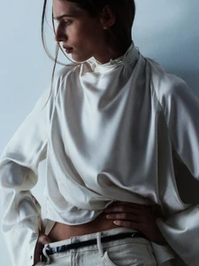 Model wearing a white satin high-neck top and light-colored trousers against a plain studio background.