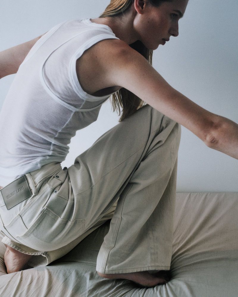 Model in white ribbed tank top and light trousers kneeling on textured bedding