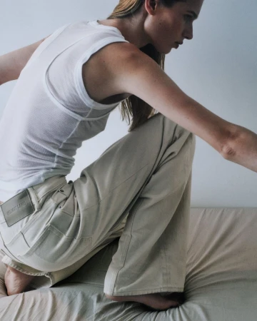 Model in white ribbed tank top and light trousers kneeling on textured bedding