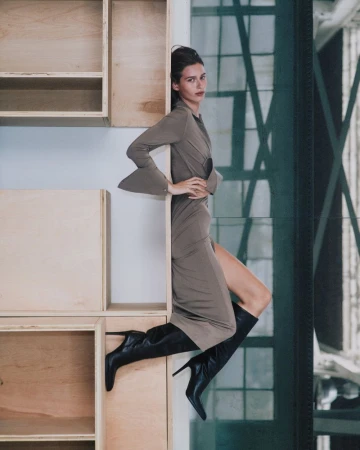 Model wearing a grey dress and black boots posing against wooden shelving for Helmut Lang campaign