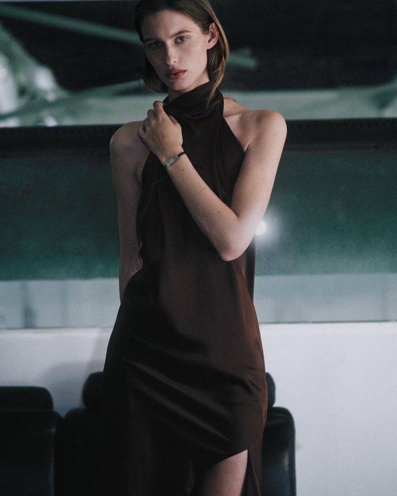Model in a dark brown halterneck dress posing against a contemporary interior background for Helmut Lang campaign.