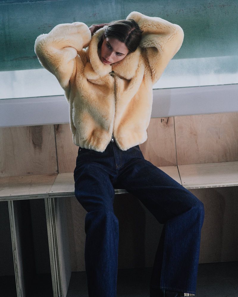 Model wearing a fluffy yellow jacket and dark denim jeans seated against a wooden panel wall for Helmut Lang