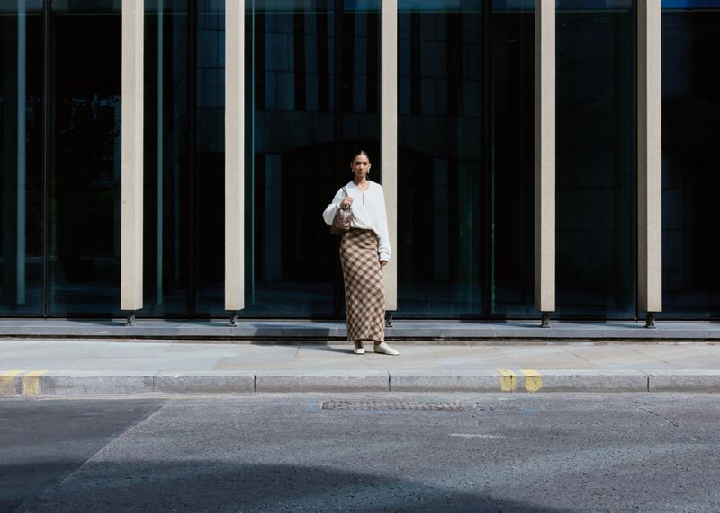 Woman standing on a city sidewalk in a white top and plaid maxi skirt in front of a modern glass building