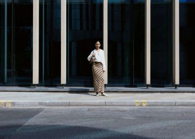 Woman standing on a city sidewalk in a white top and plaid maxi skirt in front of a modern glass building