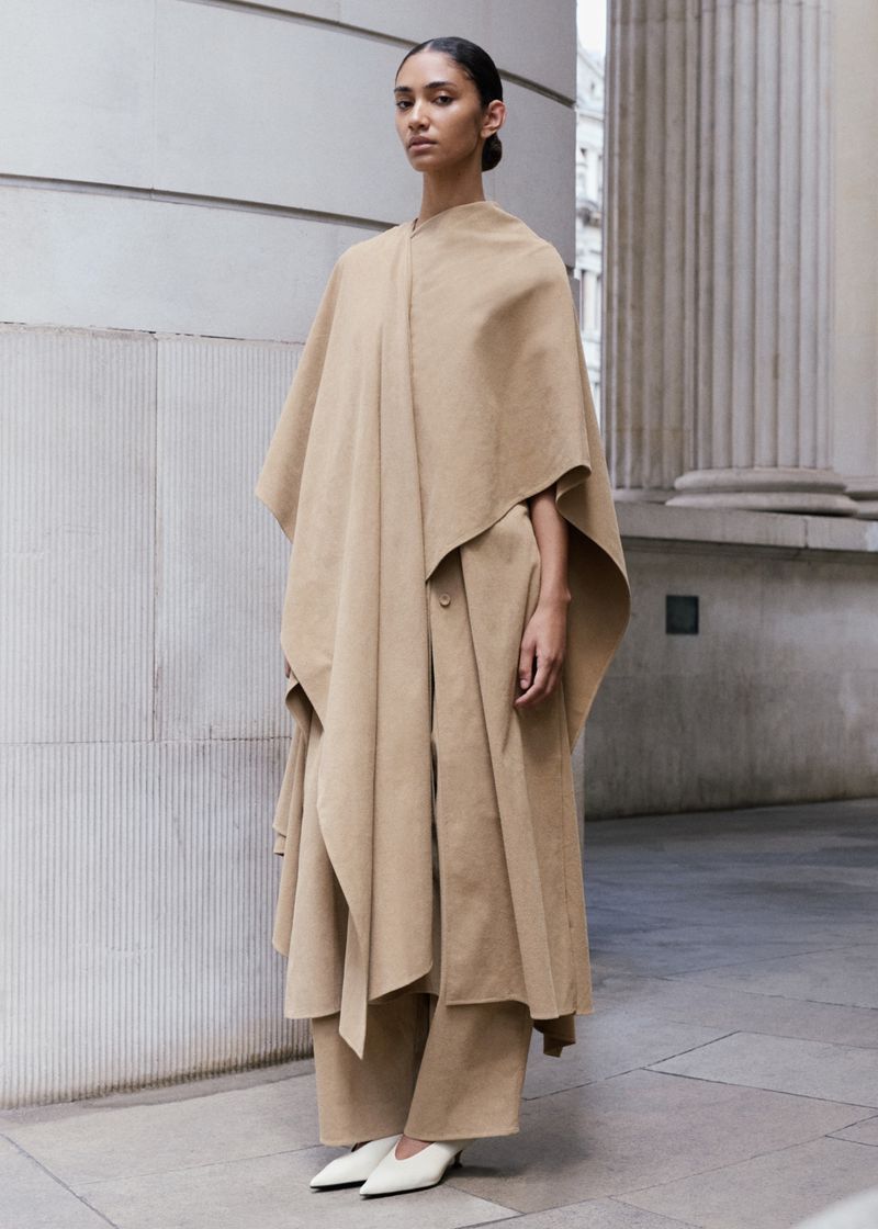 Model in camel-colored cape and matching trousers posing outdoors against stone architectural columns