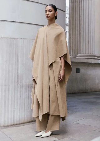 Model in camel-colored cape and matching trousers posing outdoors against stone architectural columns