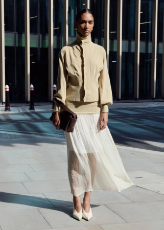 Model standing outdoors on paved ground wearing a cream high-neck top and white pleated skirt while holding a dark handbag.