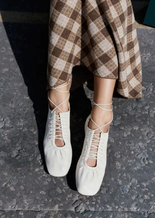 Close-up of a person wearing white lace-up leather flats and a brown gingham patterned skirt outdoors on stone pavement.