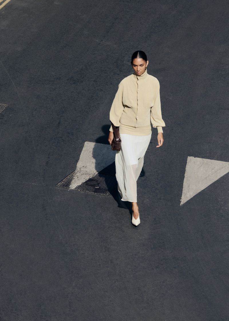 Model walking on dark asphalt road wearing cream blouse and white skirt holding a dark handbag