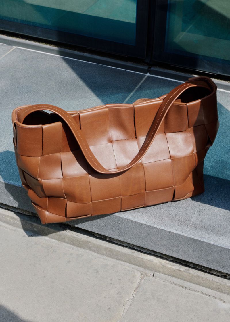 Brown leather woven shoulder bag resting on grey stone steps in bright natural light.