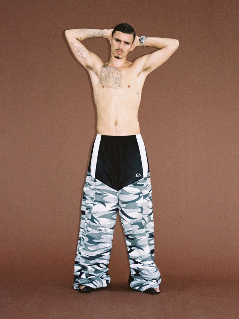 Man with tattoos stands in wide-leg camo pants against a brown backdrop, arms behind his head, displaying a watch and casual style.