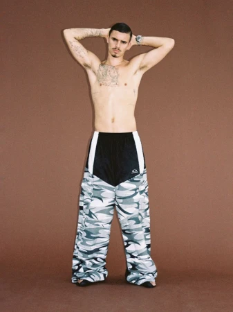 Man with tattoos stands in wide-leg camo pants against a brown backdrop, arms behind his head, displaying a watch and casual style.