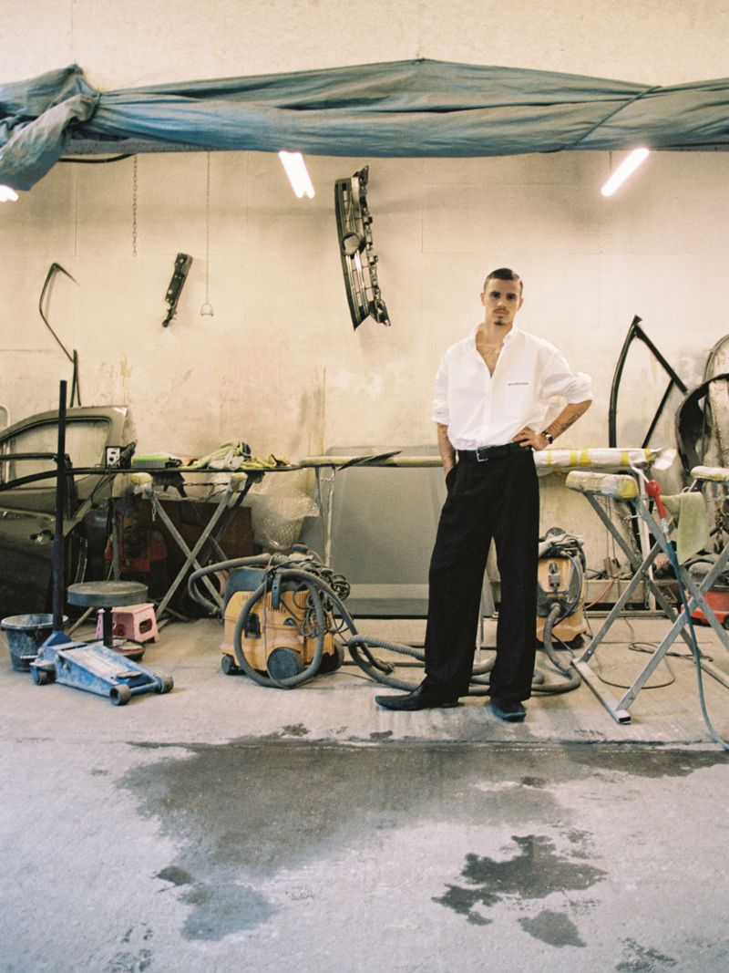 Adult male in an open white shirt and dark pants standing in a car repair shop with tools and car parts visible.