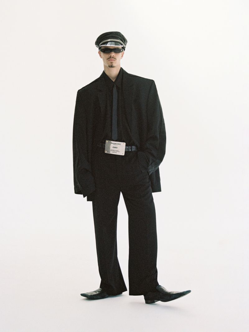Stylish adult male in a black suit, tie, and hat, wearing sunglasses, standing against a bright white backdrop in a fashion forward pose.