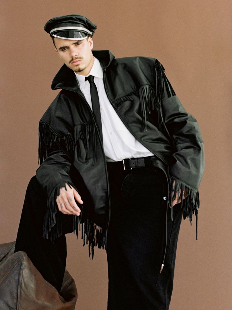Adult male fashion model in a black fringe jacket, white shirt, black tie, and leather cap poses in a studio with a brown backdrop.