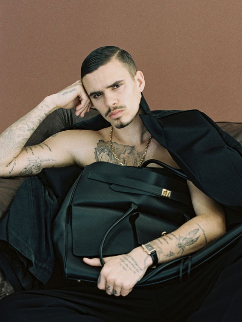 Portrait of a young adult male with tattoos, gold jewelry, and a black bag in an indoor setting against a brown backdrop.