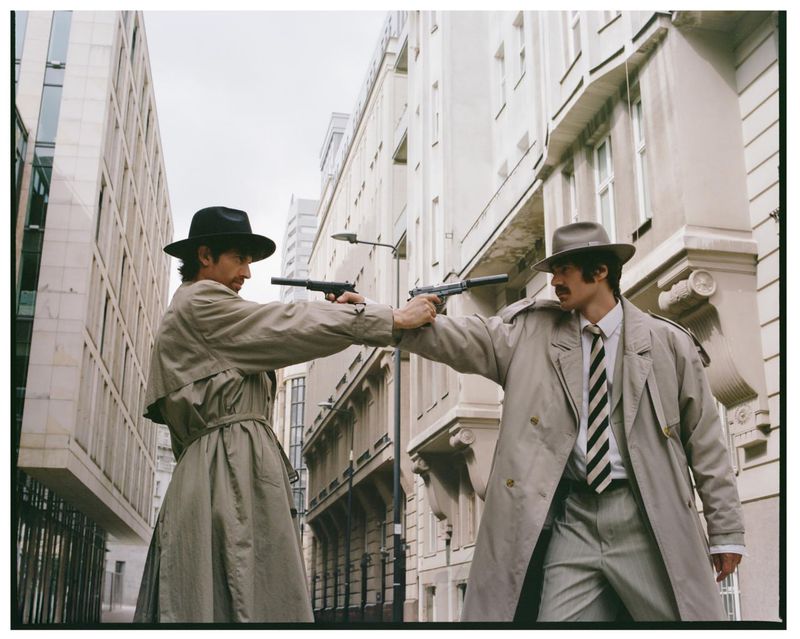 Two men in trench coats and fedoras standing in an urban street holding silenced pistols pointed at each other